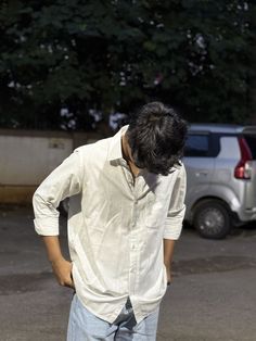 Person wearing a light-colored shirt and jeans standing outdoors with a vehicle and trees in the background.