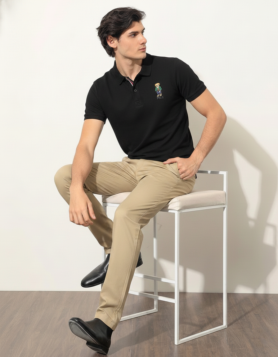 Man sitting on a stool wearing a black polo shirt and beige pants against a white wall.