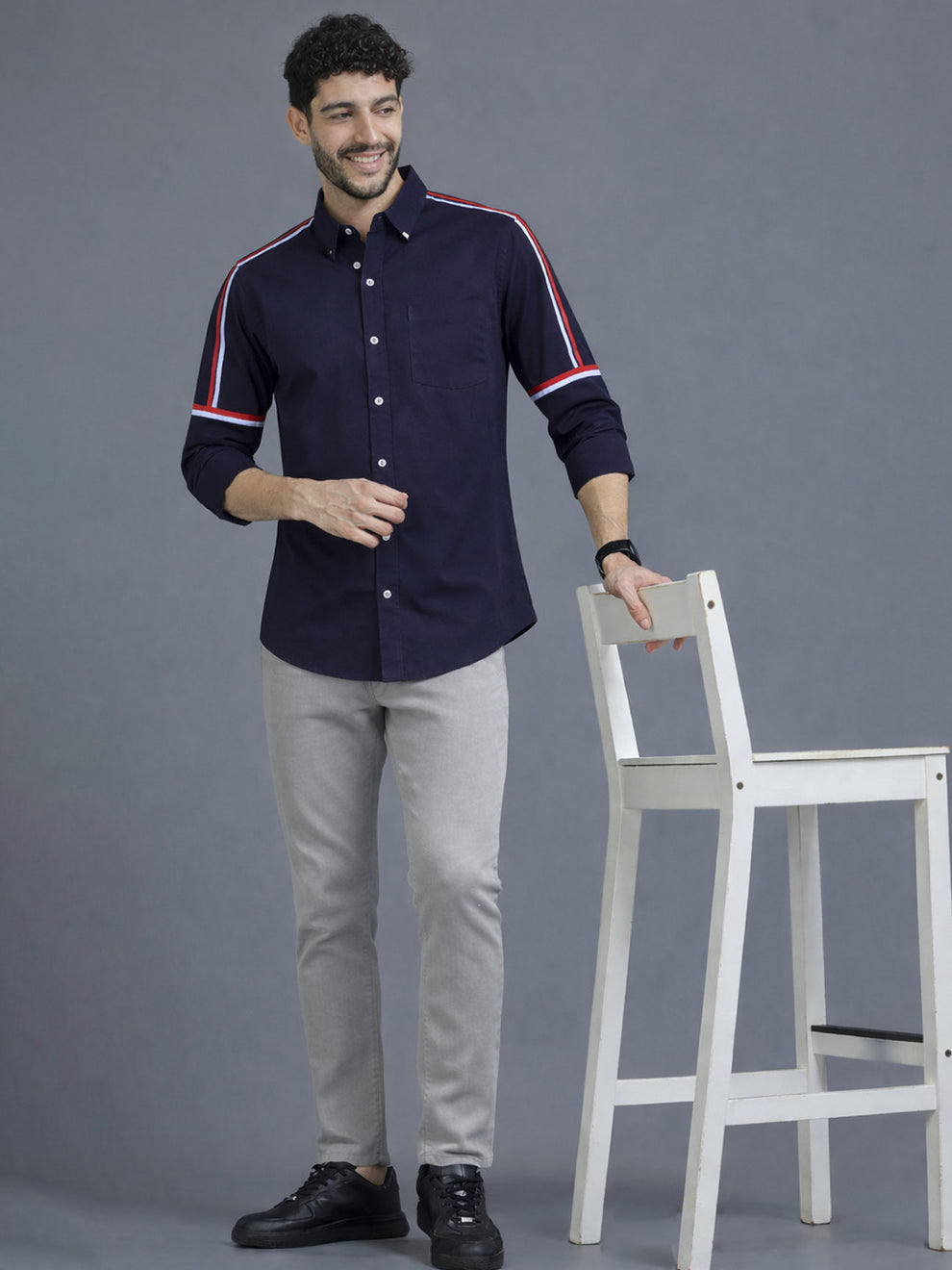 Man wearing a navy shirt with white stripes standing next to a white chair against a gray background