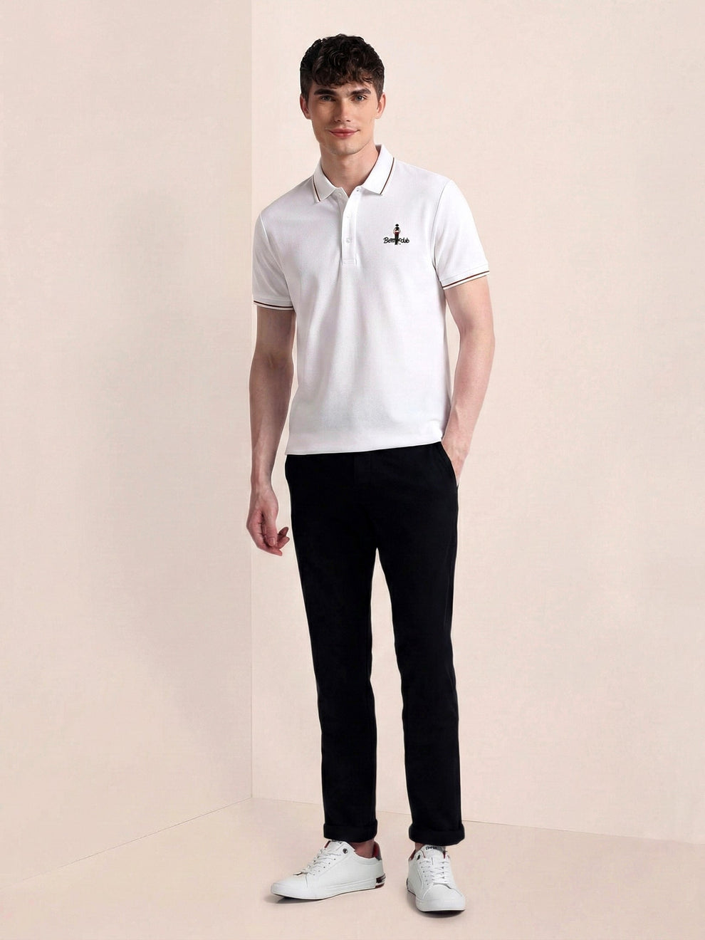 Man wearing a white polo shirt with a logo and black pants on a beige background