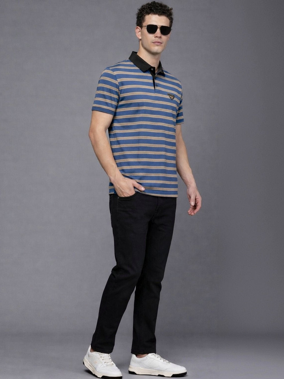 Man wearing a blue and gray striped polo shirt with black collar, black pants, and white sneakers on a gray background.