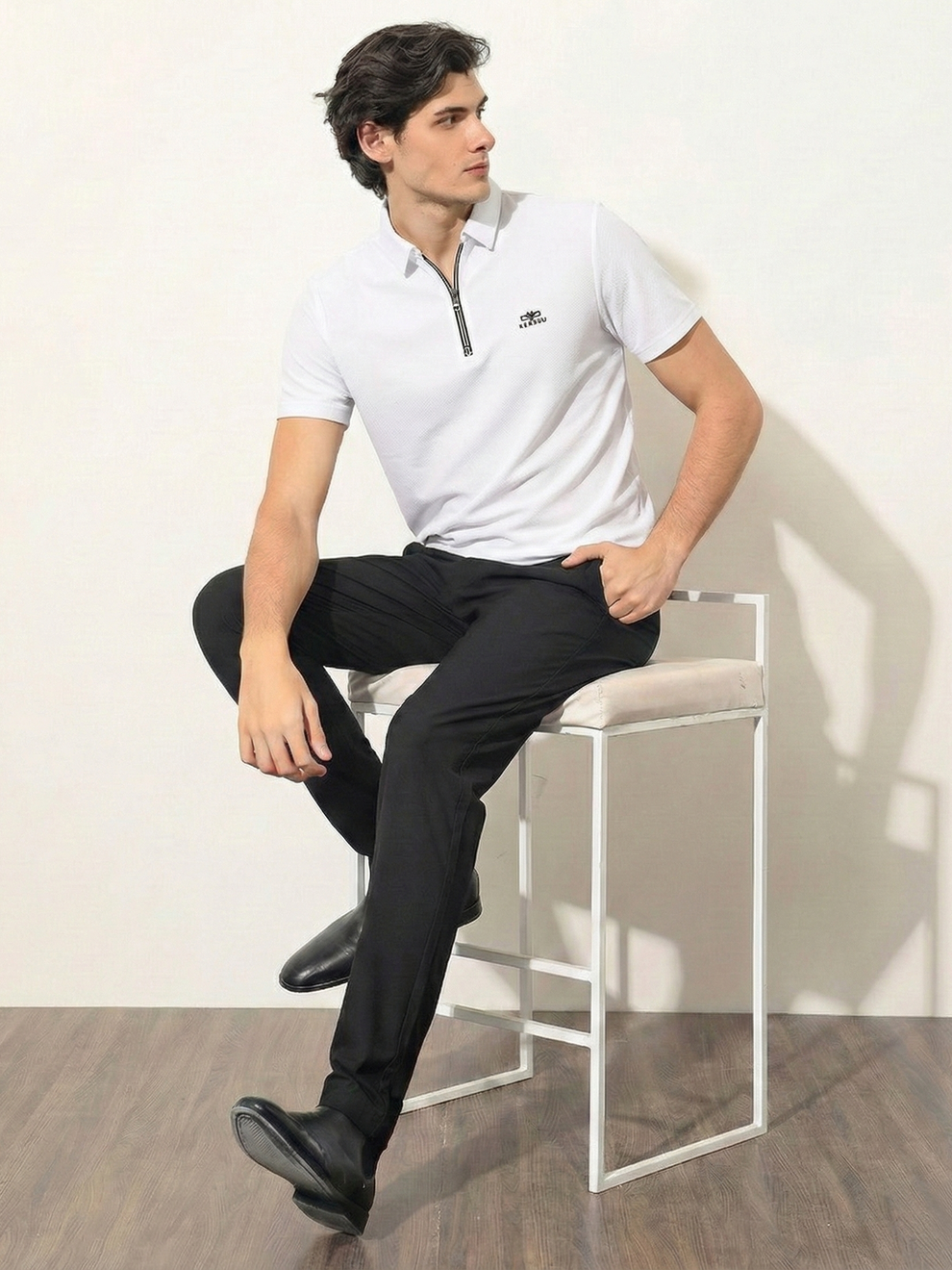 Man sitting on a stool wearing a white polo shirt and black pants against a plain background