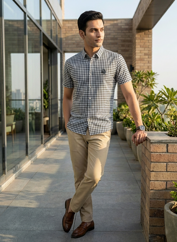 Man in a checkered shirt and beige pants standing on a rooftop with plants and cityscape in the background