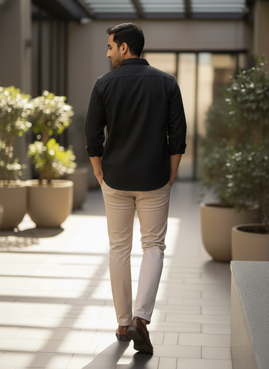 Man in black shirt and beige pants walking outdoors with plants and a building in the background