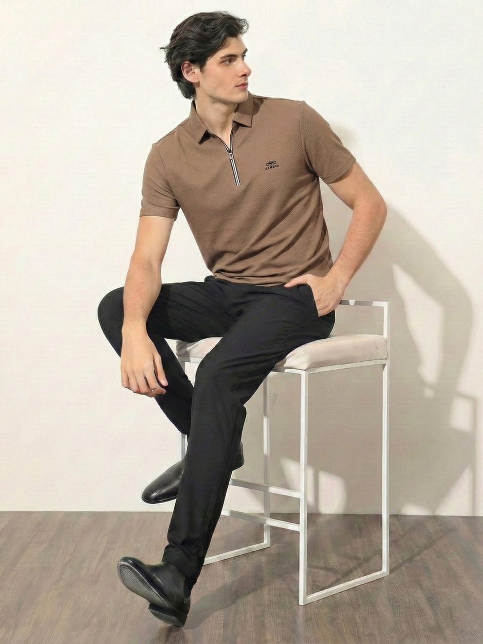 Man sitting on a chair wearing a brown polo shirt and black pants against a white wall.
