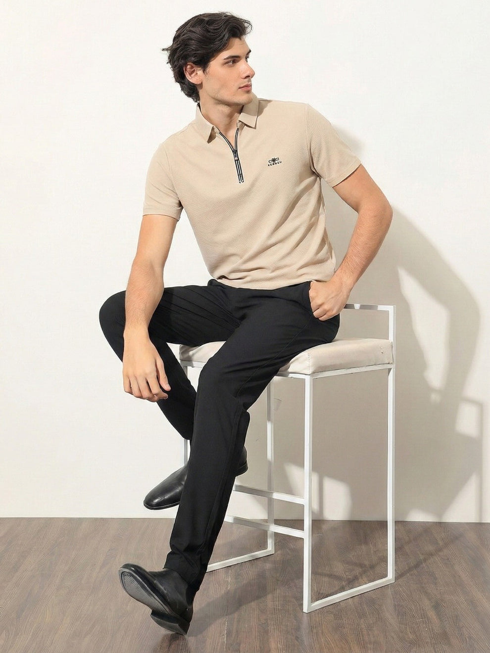 Man sitting on a stool wearing a beige polo shirt and black pants against a white wall.