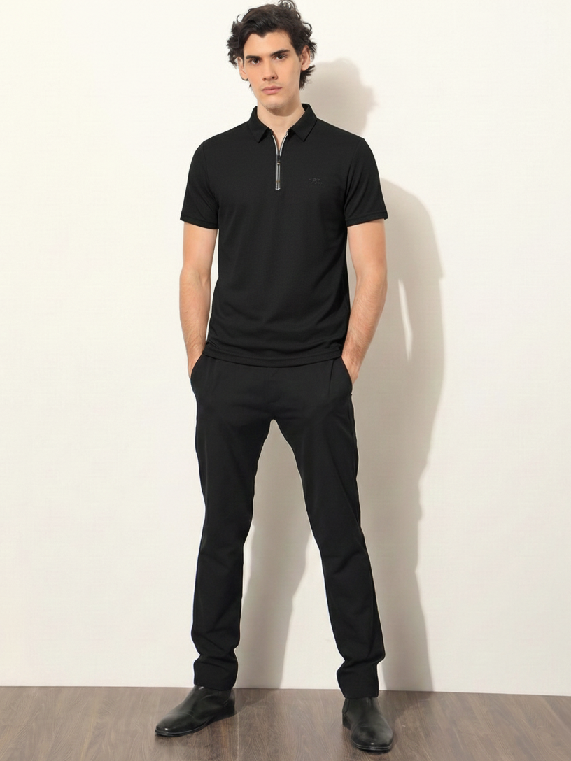 Man wearing a black polo shirt and pants against a white wall.