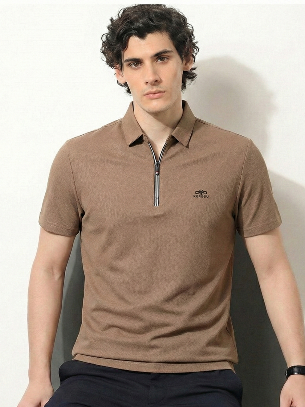 Man wearing a brown polo shirt with a zipper sitting against a white wall.