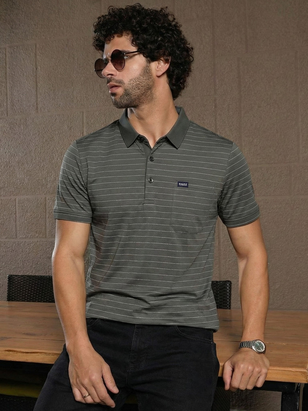 Man wearing a striped polo shirt with a brand logo, sitting at a table against a textured wall.