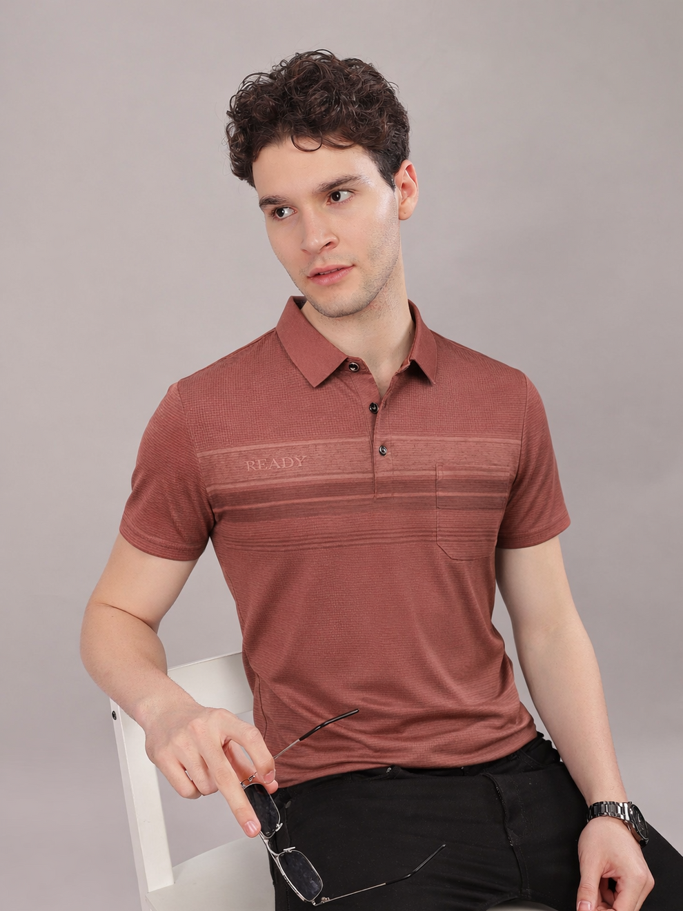 Man wearing a red polo shirt with a brand logo, sitting on a white chair against a gray background.