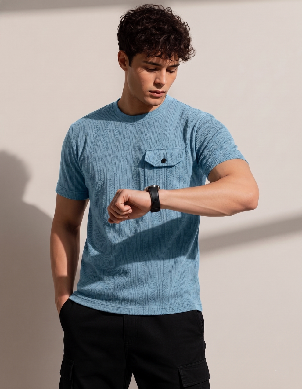 Man wearing a blue textured shirt and black pants, looking at his watch against a neutral background