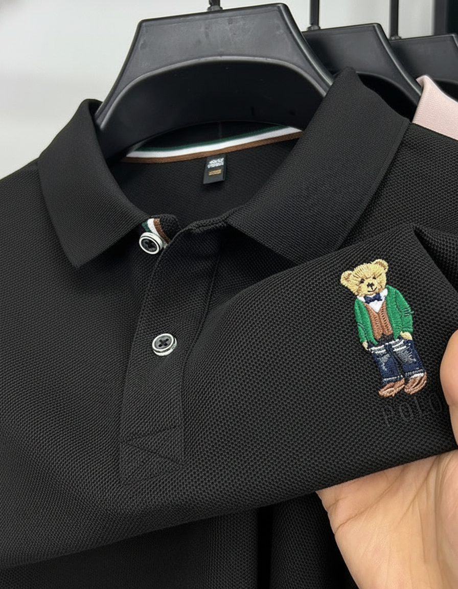 Black polo shirt with embroidered bear design held by a hand against a neutral background