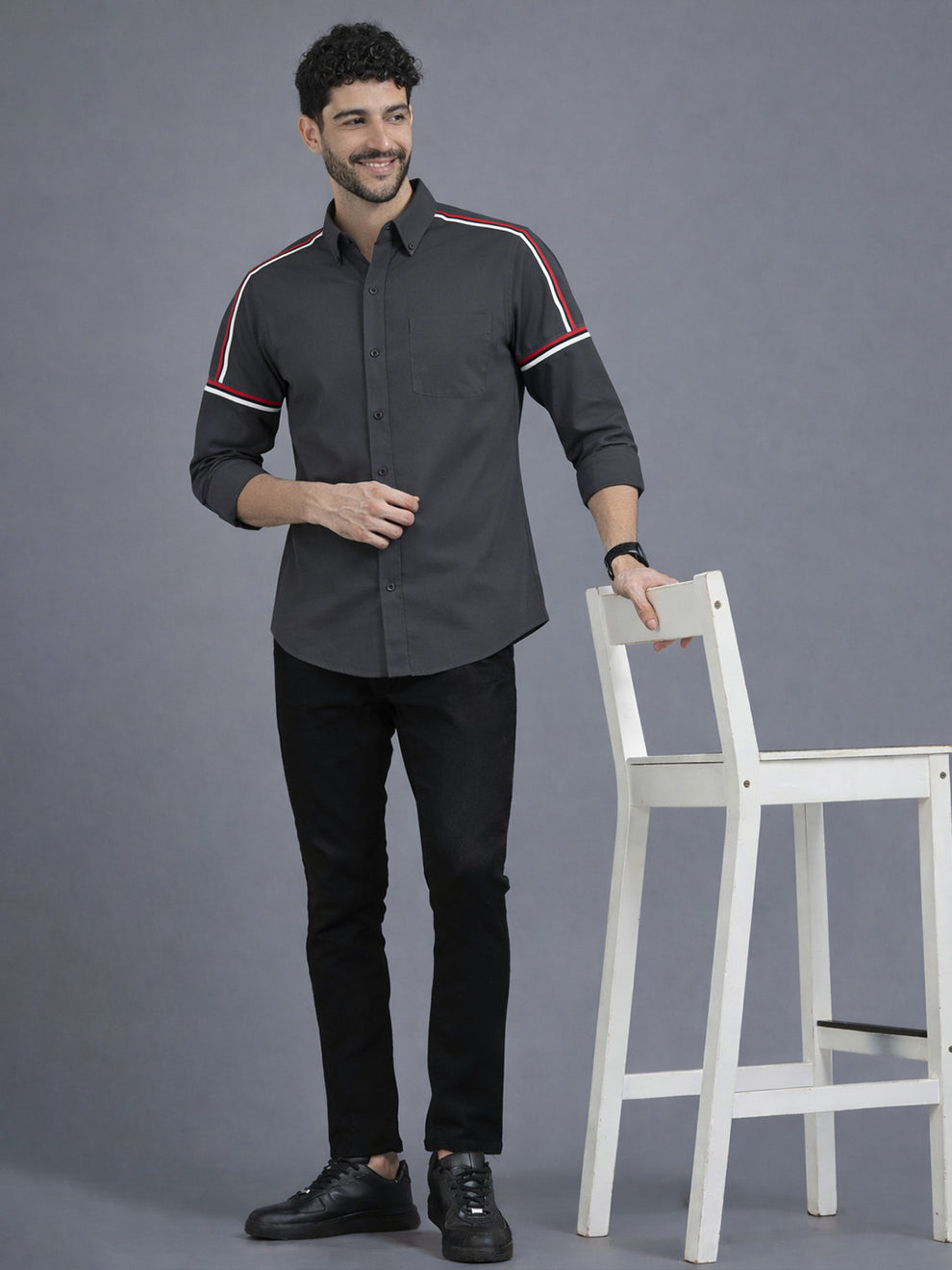 Man wearing a dark gray shirt with red and white accents, standing next to a white chair on a gray background