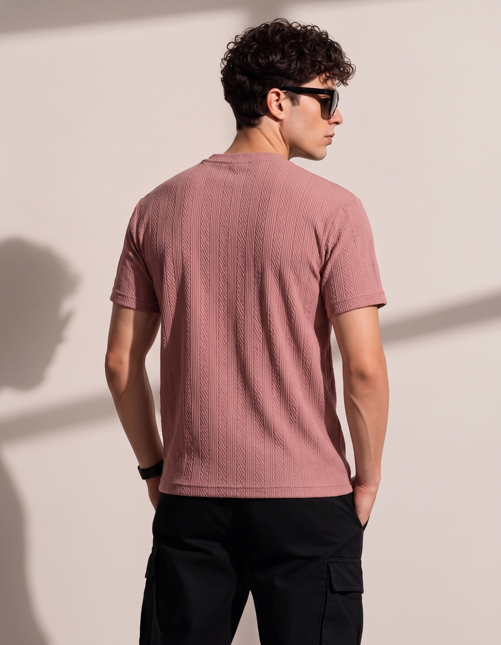 Man wearing a pink textured t-shirt and black sunglasses against a beige wall
