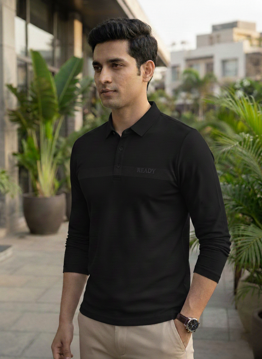 Man wearing a black polo shirt with a brand logo, standing outdoors with plants and buildings in the background.