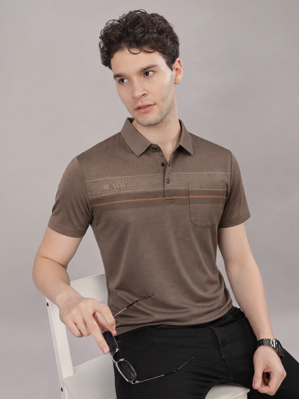 Man wearing a brown polo shirt with a visible brand logo, sitting on a white chair against a gray background.