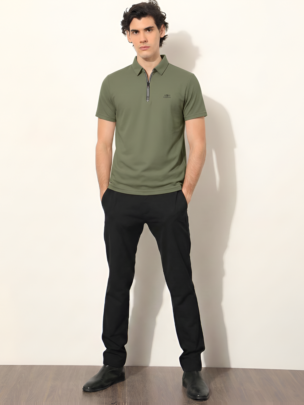 Man wearing a green polo shirt and black pants against a white wall.