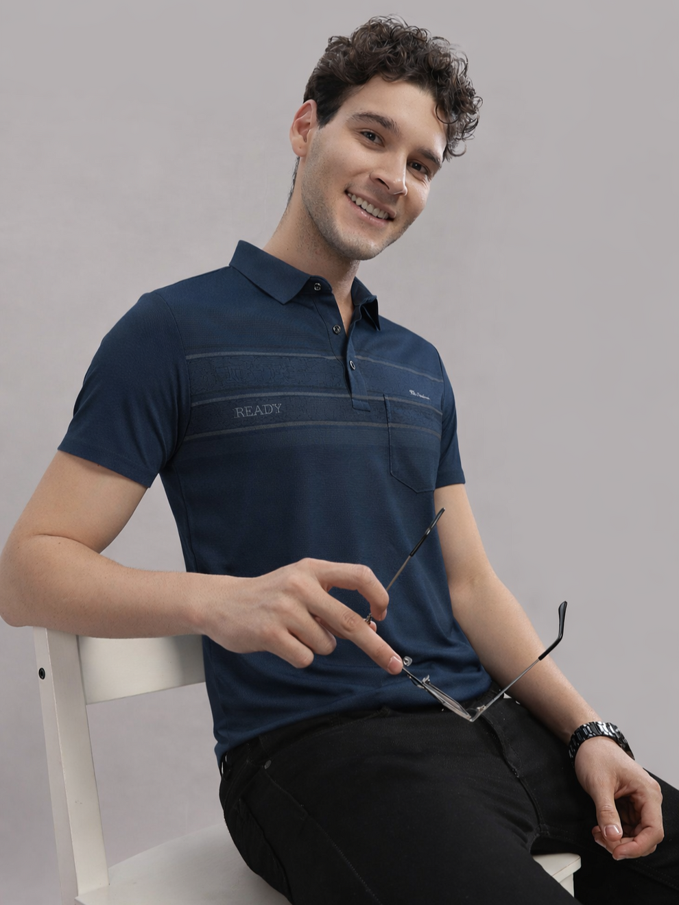 Man wearing a navy blue polo shirt sitting on a white chair against a gray background