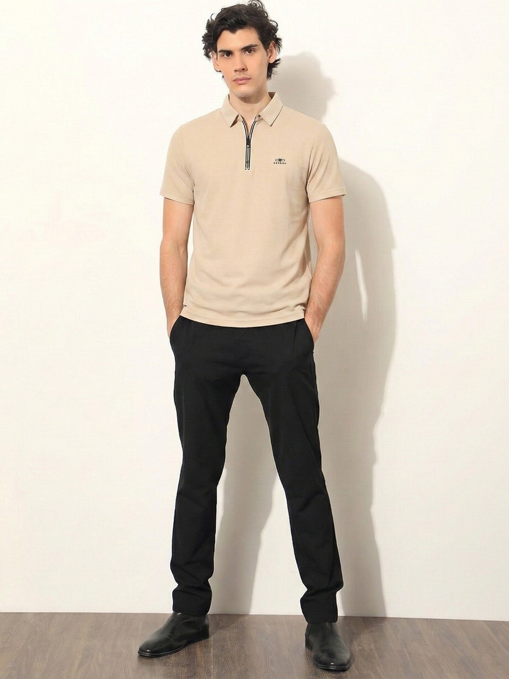 Man wearing a beige polo shirt and black pants against a white wall.
